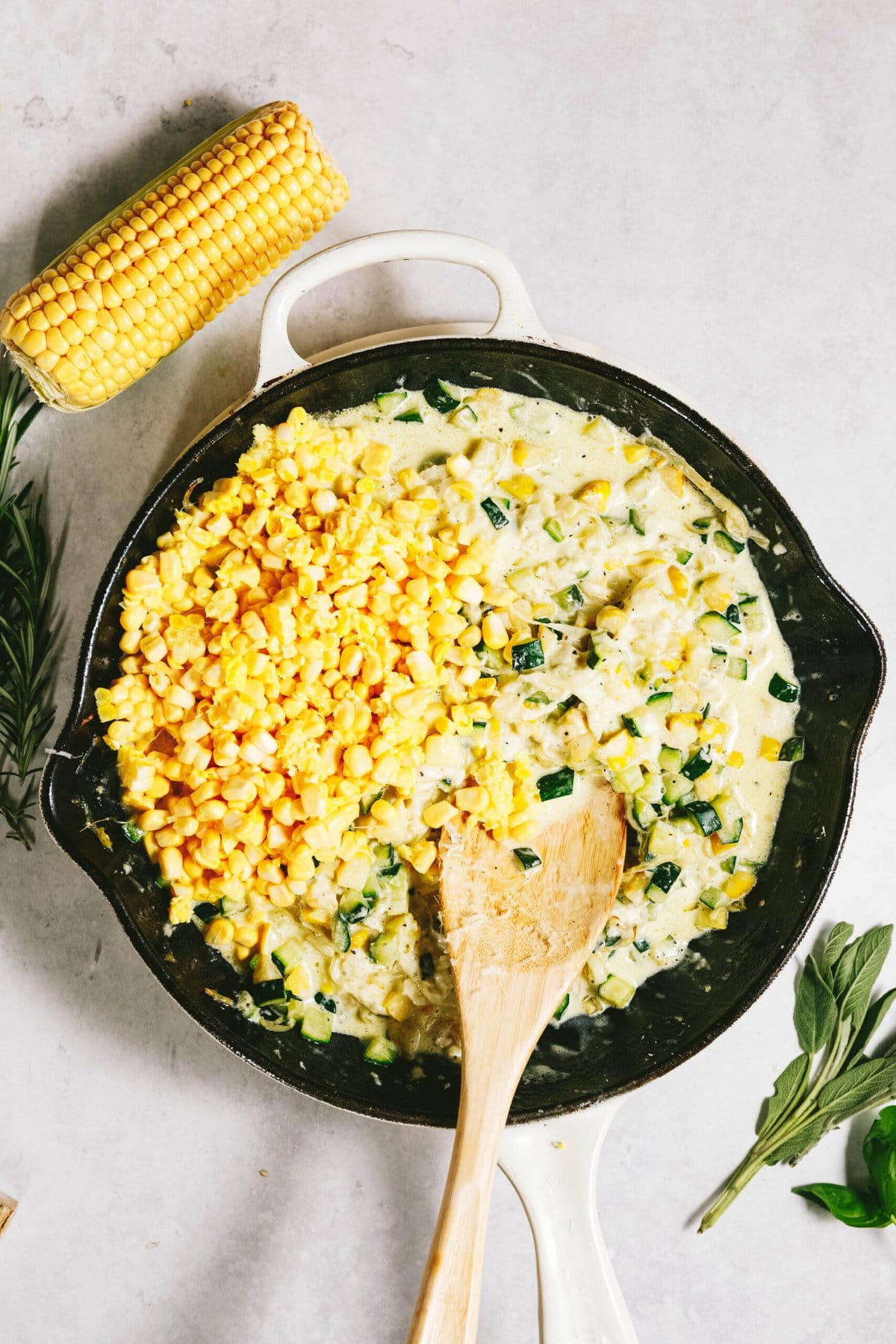 A skillet with a creamy corn, herbs, and zucchini mixture evokes the essence of summer squash. A wooden spoon rests in the pan, alongside a cob of corn and fresh herbs nearby, blending seasonal flavors like a delightful summer squash pasta dish.