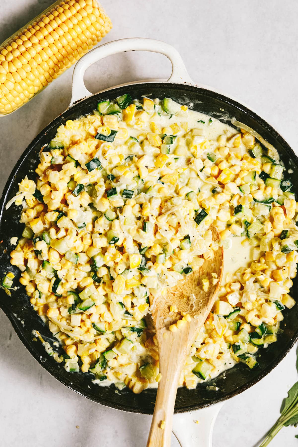 A skillet brims with a creamy blend of corn, zucchini, and summer squash. A wooden spoon rests inside the skillet, and a corn cob lies nearby on a light surface.