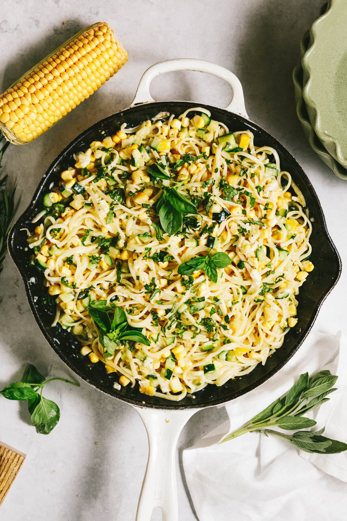 A skillet of summer squash pasta mixed with corn and herbs, garnished with basil leaves, sits invitingly next to a cob of corn and green plate.