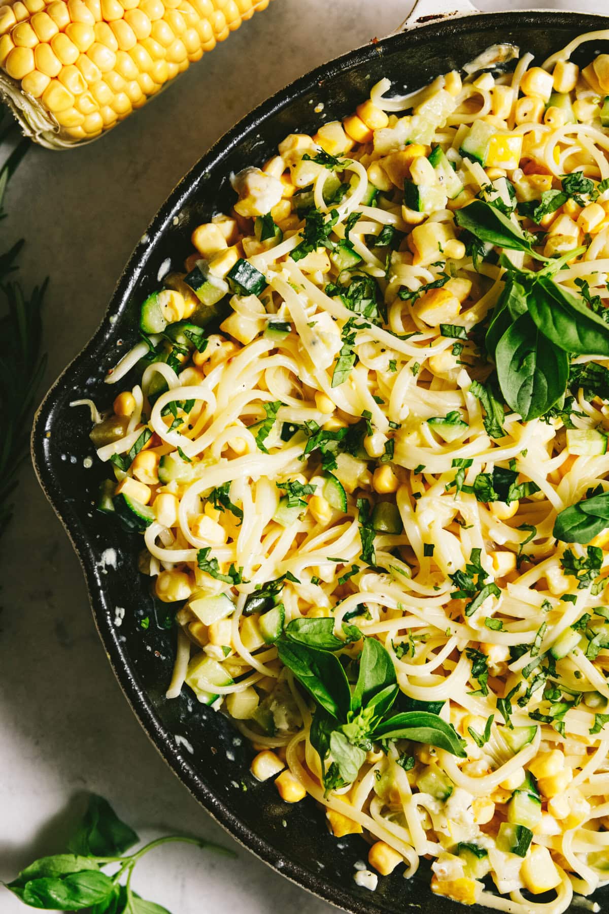 Pasta with summer squash, corn, basil, and herbs in a skillet, garnished with fresh greens. Corn cob and basil leaves are visible beside the skillet.