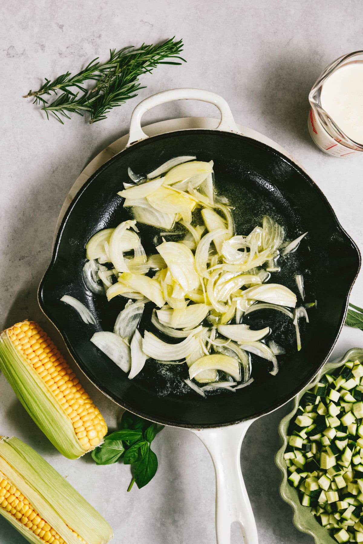 A black skillet with sliced onions rests on a gray surface, surrounded by fresh corn, zucchini, and rosemary. A cup of cream hints at the richness to come in this summer squash pasta delight.