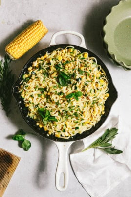 A skillet with summer squash pasta, corn, and fresh herbs sits on a table next to a cob of corn, plates, and a white cloth.