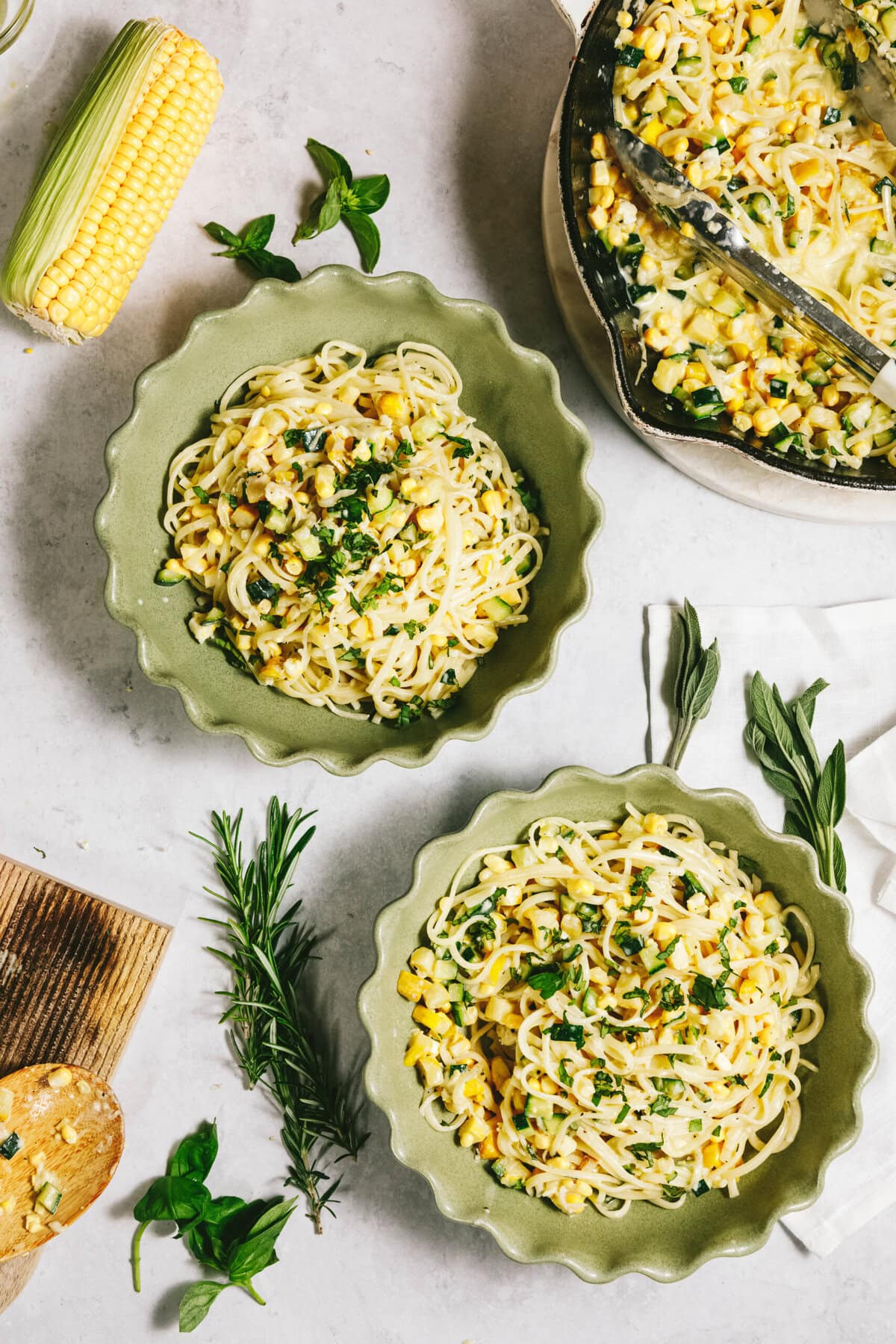 Two bowls of summer squash pasta with corn and herbs are placed on a table. A skillet with additional pasta, an ear of corn, and fresh herbs are also visible.