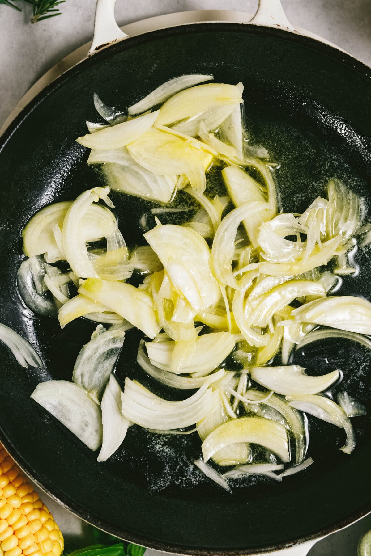 Sliced onions sauté in a black skillet, with summer squash adding a fresh touch and a corn cob partially visible at the bottom left.