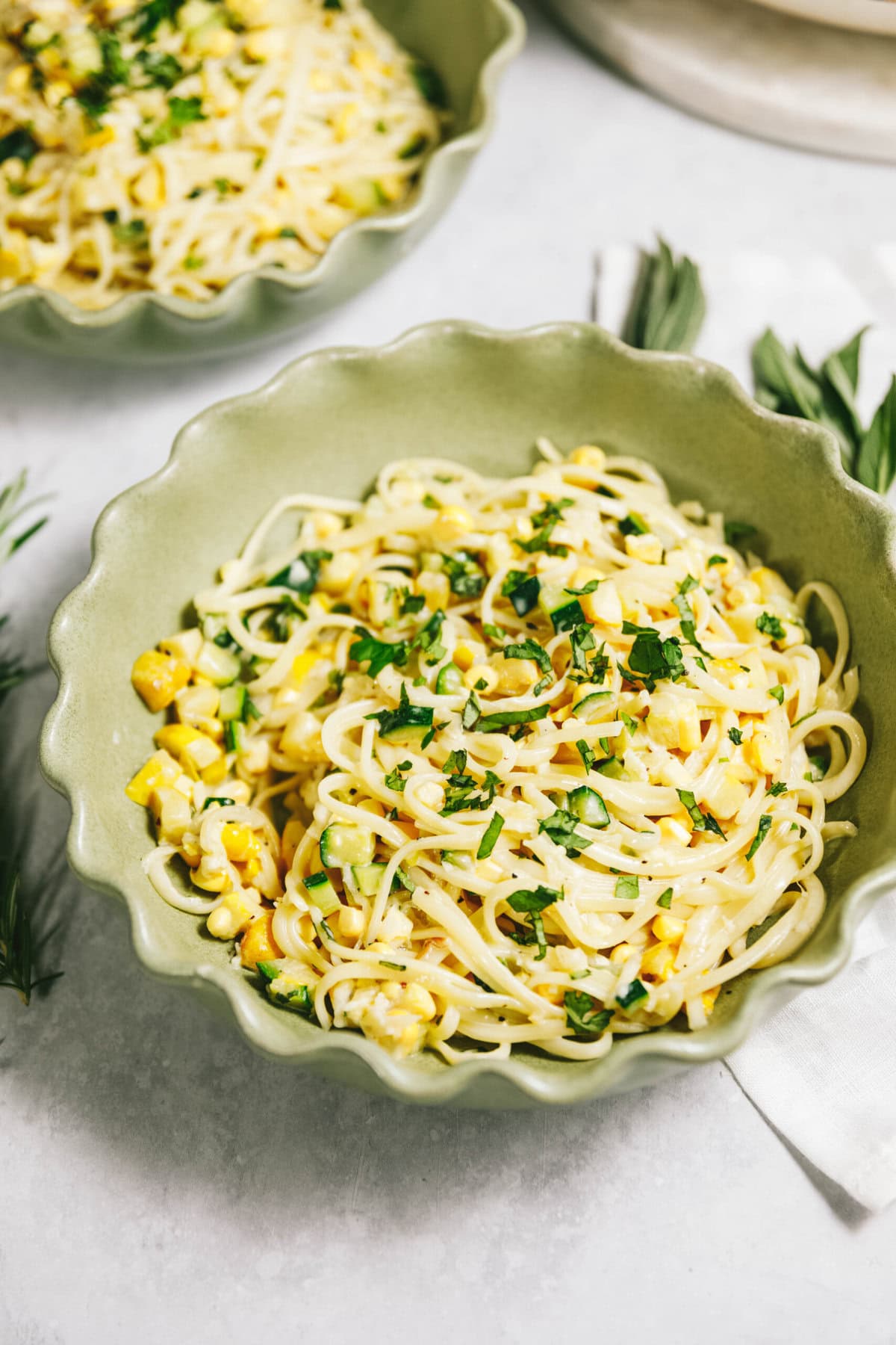 Two bowls of summer squash pasta with corn and herbs, served in green scalloped bowls on a light-colored surface.