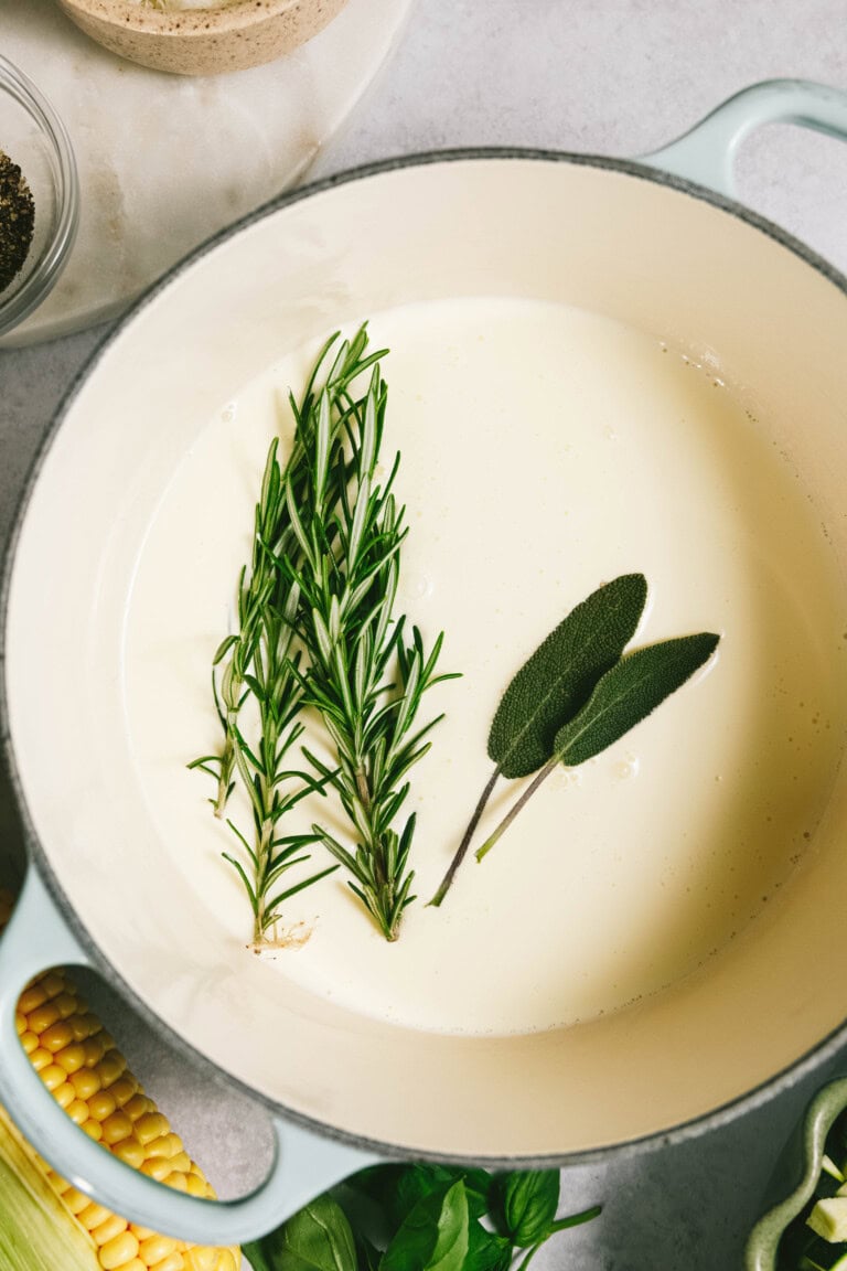 A pot filled with cream, rosemary, and sage bubbles gently alongside a vibrant summer squash pasta. Corn and basil are placed nearby, waiting to enhance the delightful medley of flavors.