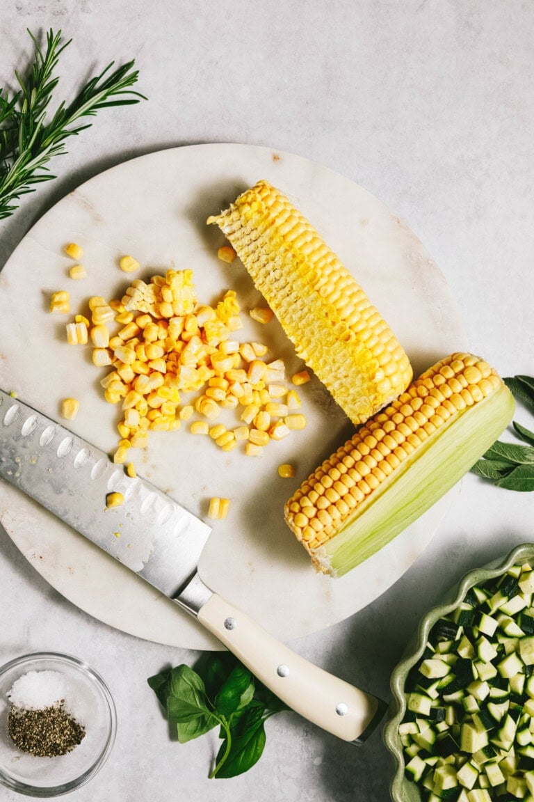 Chopped corn on a cob and fresh herbs adorn a cutting board with a knife, joined by summer squash and a bowl of diced vegetables, ready to complement your favorite pasta dish.