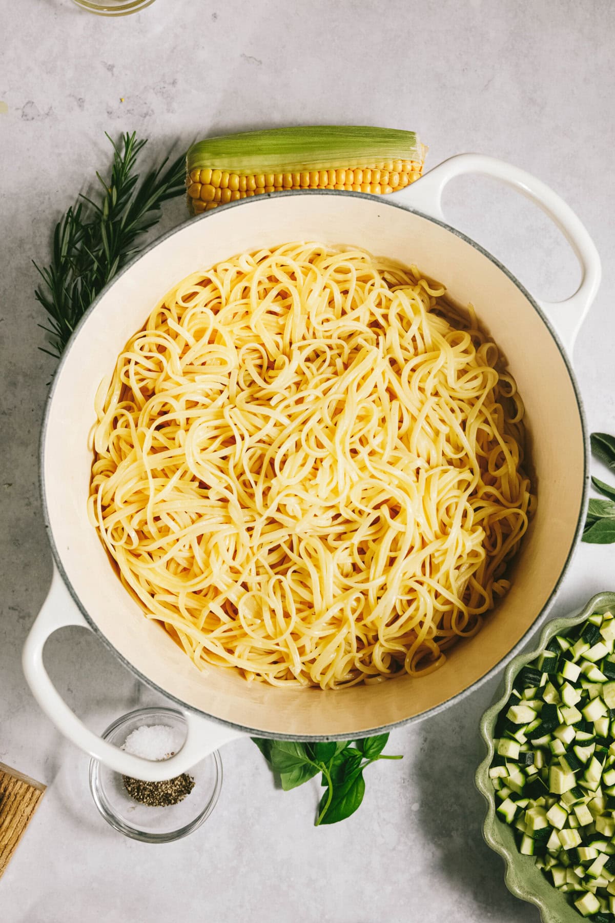 A white pot filled with summer squash pasta is surrounded by herbs, a corn cob, a small bowl of pepper, and chopped zucchini on a gray surface.