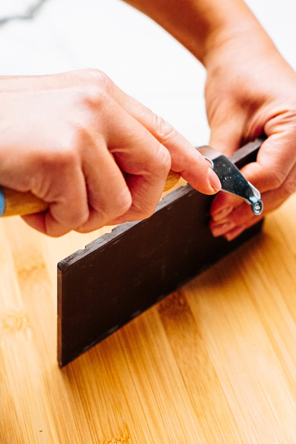 A person expertly uses a potato peeler to create delicate chocolate curls for cake on a wooden surface.