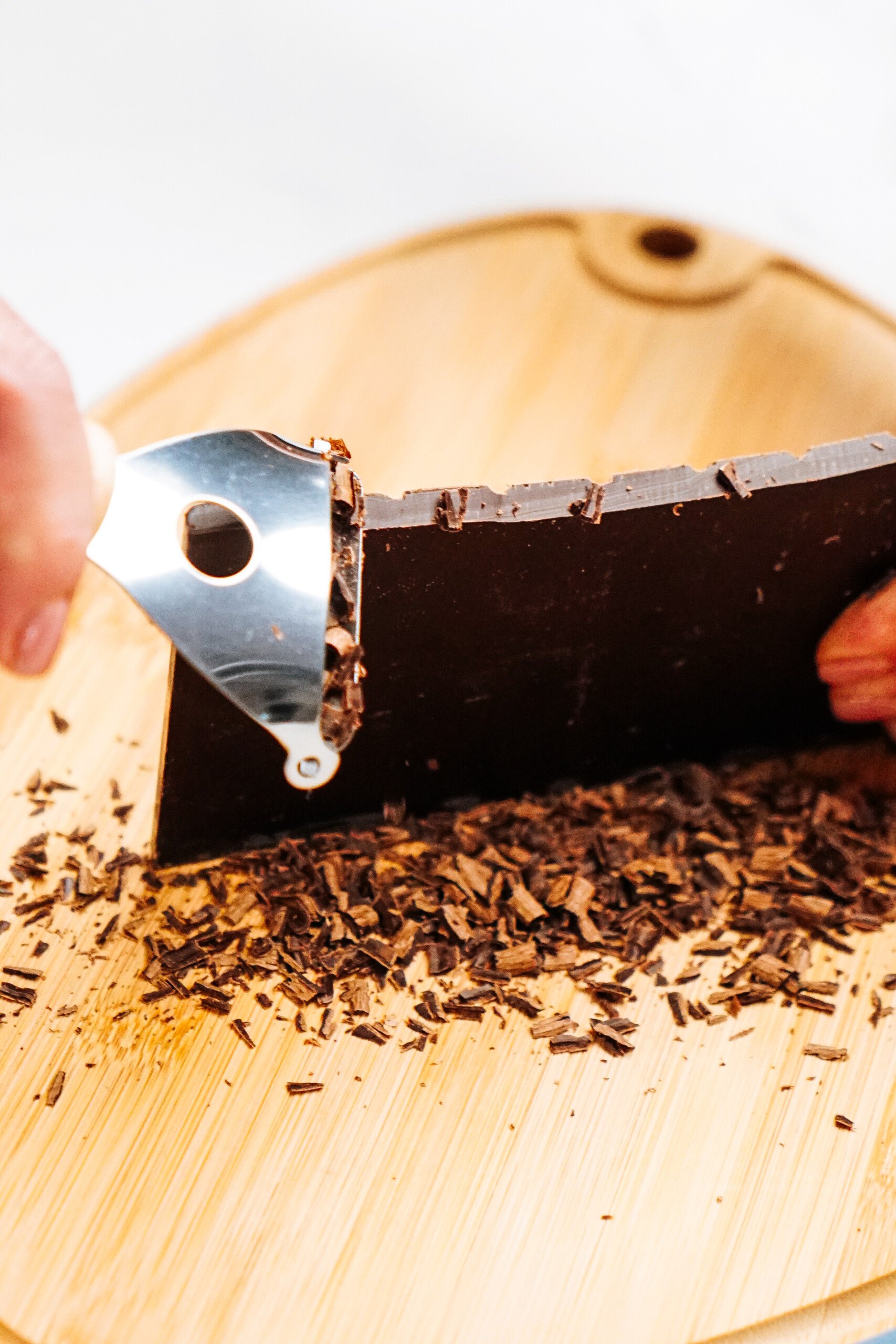 Person chopping dark chocolate on a wooden board with a scraper tool, creating small chocolate shavings.