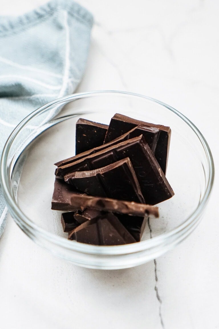 A glass bowl containing several chunks of dark chocolate and delicate chocolate curls for cake, placed on a light surface near a folded blue cloth.