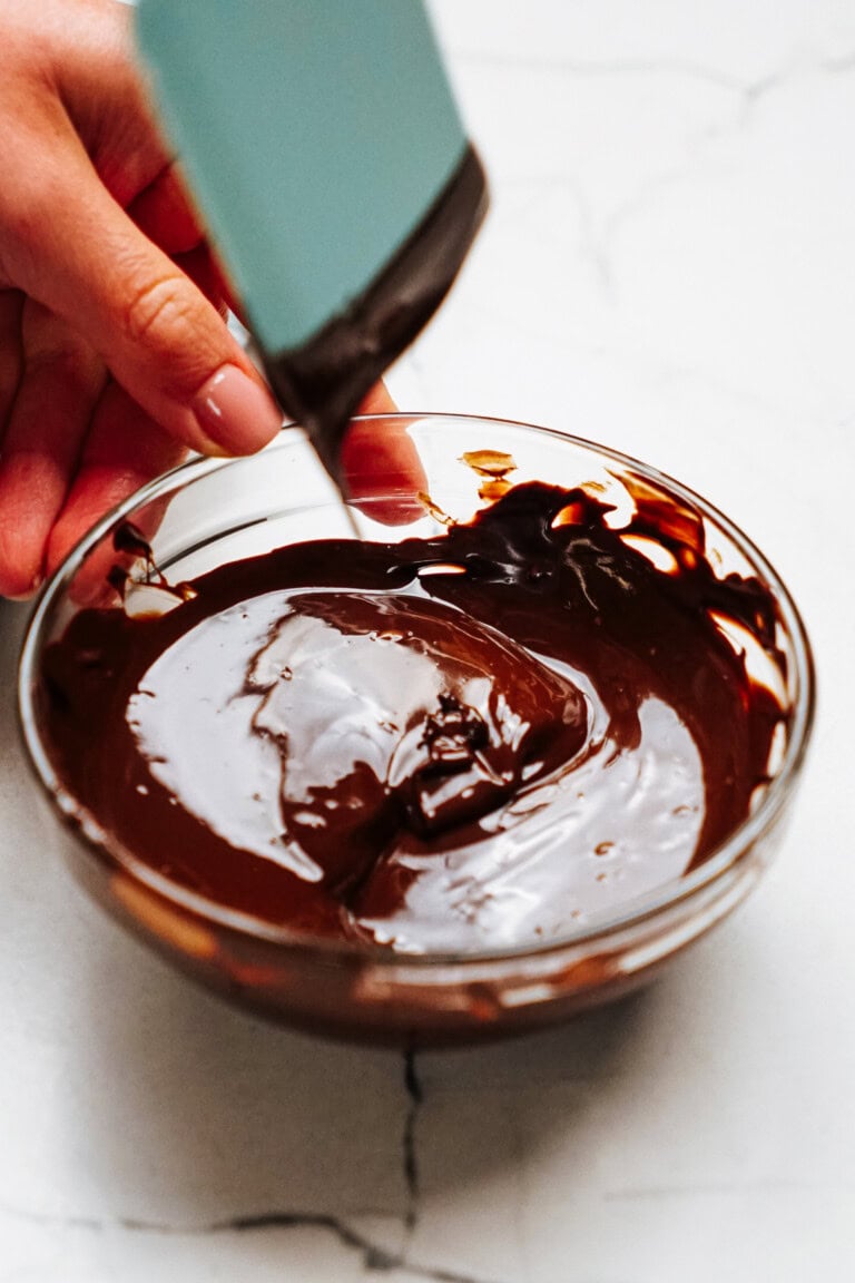 A hand using a blue spatula to stir melted chocolate in a glass bowl on a marble countertop, preparing the smooth base for delicate chocolate curls for cake.