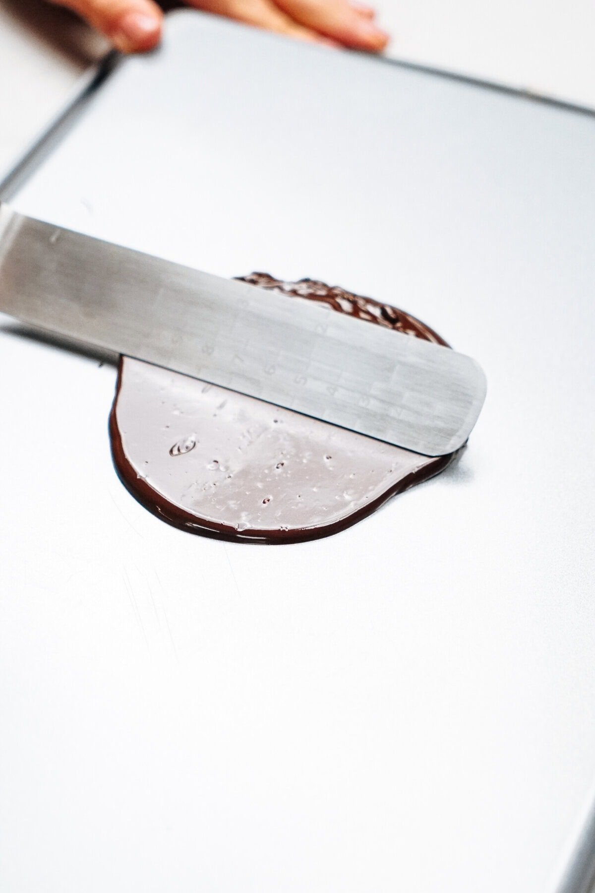 A metal spatula spreads melted chocolate on a flat surface, perfecting it into smooth layers ready to create delicate chocolate curls for cake decoration.