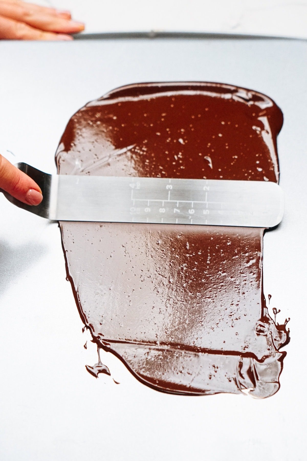 A person effortlessly spreading melted chocolate on a smooth surface with a scraper, preparing to create delicate chocolate curls for cake decorations.