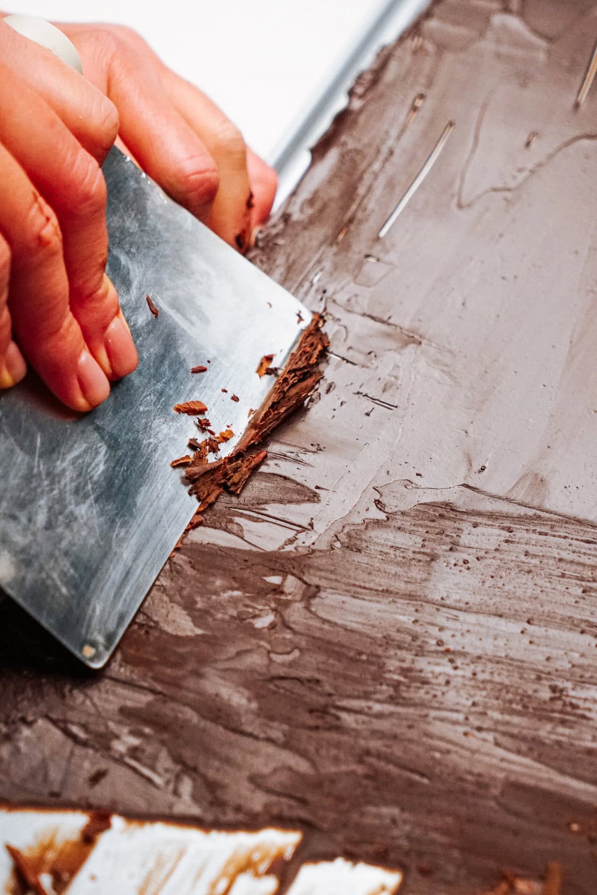 A person expertly uses a metal scraper to smooth and shape melted chocolate on a flat surface, skillfully forming delicate chocolate curls for cake decoration.