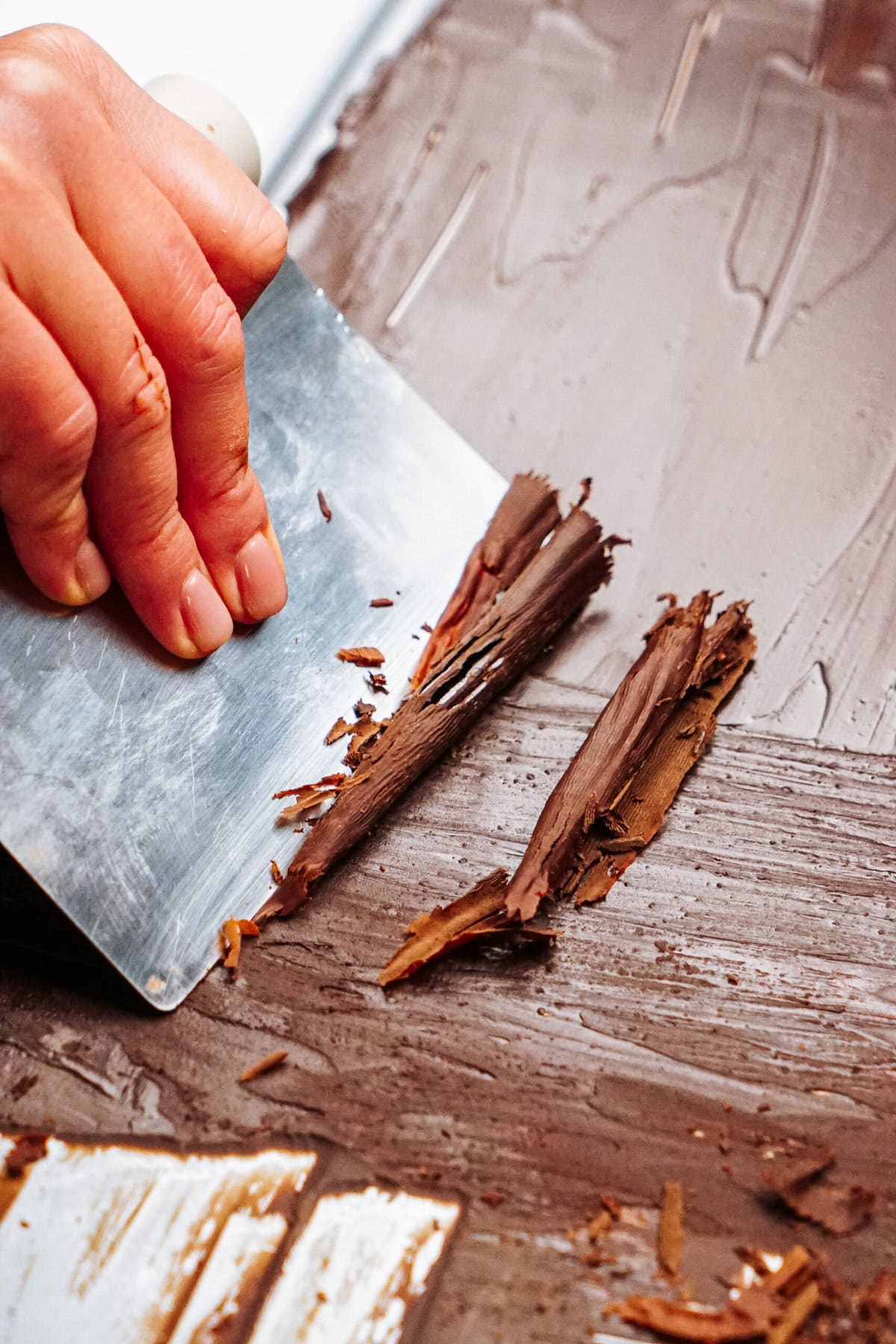 A hand expertly uses a metal scraper to create delicate chocolate curls for cake on a wooden surface.