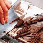 A hand expertly uses a knife to create delicate chocolate curls from a large block on a metal surface, perfect for adorning cakes.