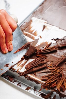 A hand expertly uses a knife to create delicate chocolate curls from a large block on a metal surface, perfect for adorning cakes.