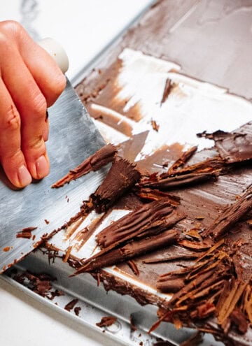 A hand expertly uses a knife to create delicate chocolate curls from a large block on a metal surface, perfect for adorning cakes.