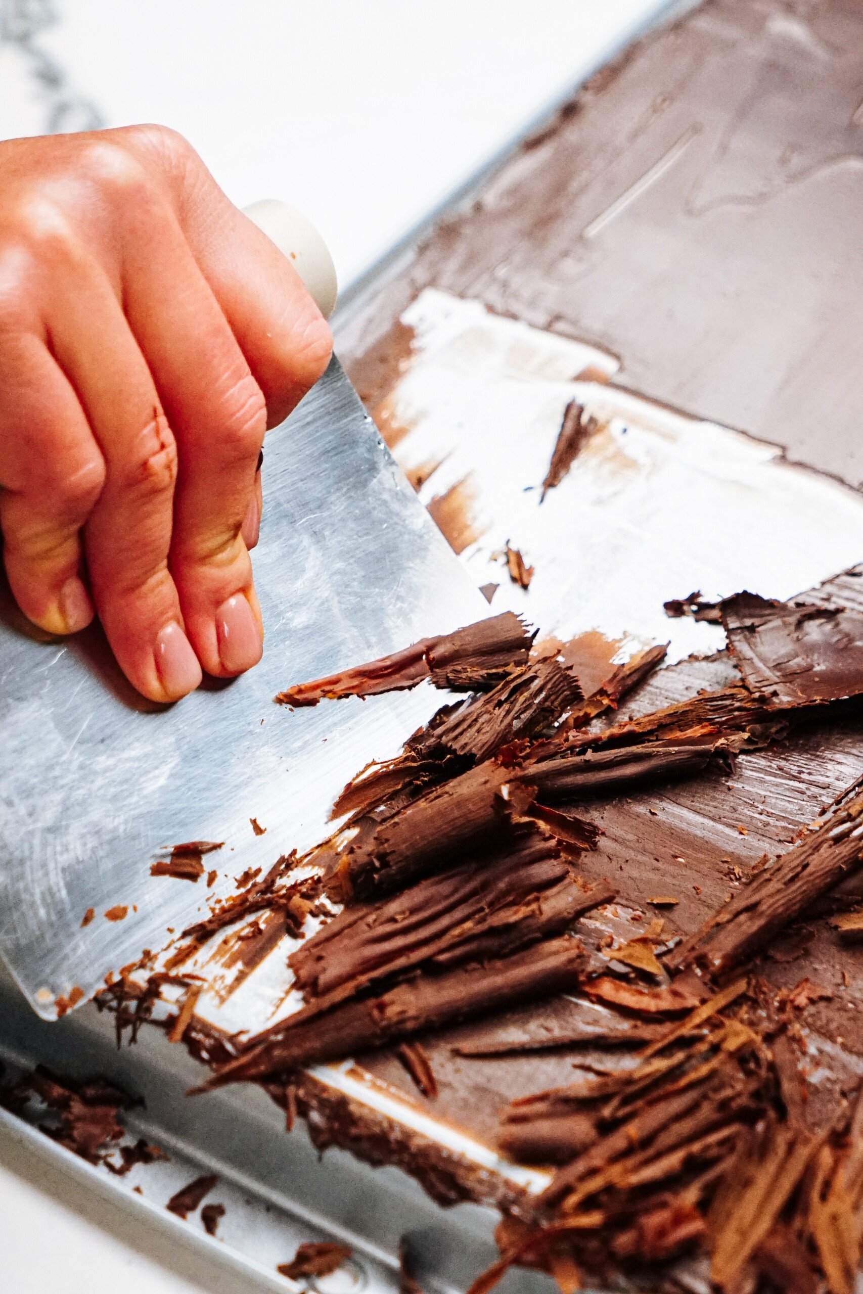 A hand uses a metal scraper to create chocolate shavings on a flat surface.