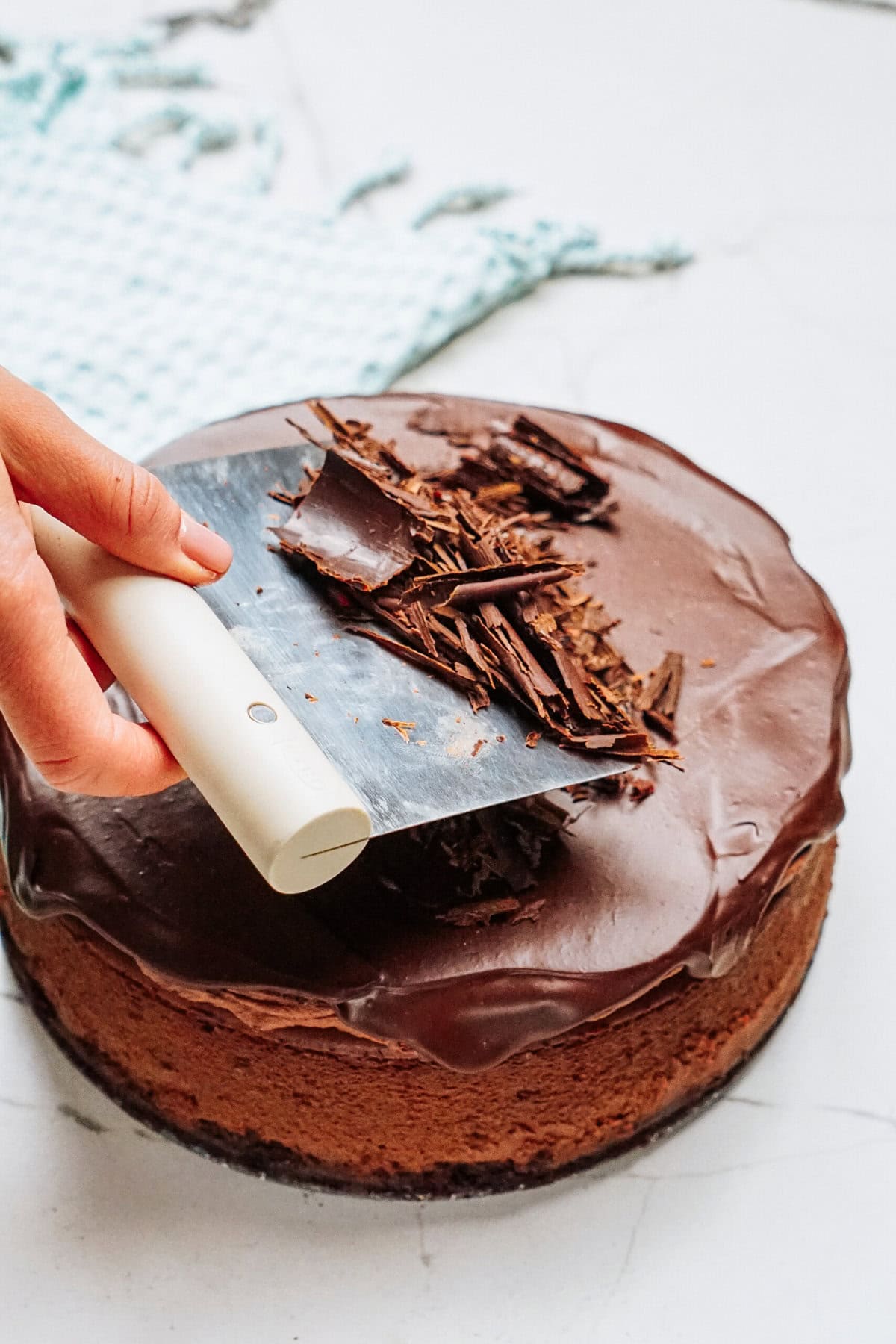 A hand uses a spatula to sprinkle chocolate curls on a chocolate cake with icing, carefully placed on a marble countertop.