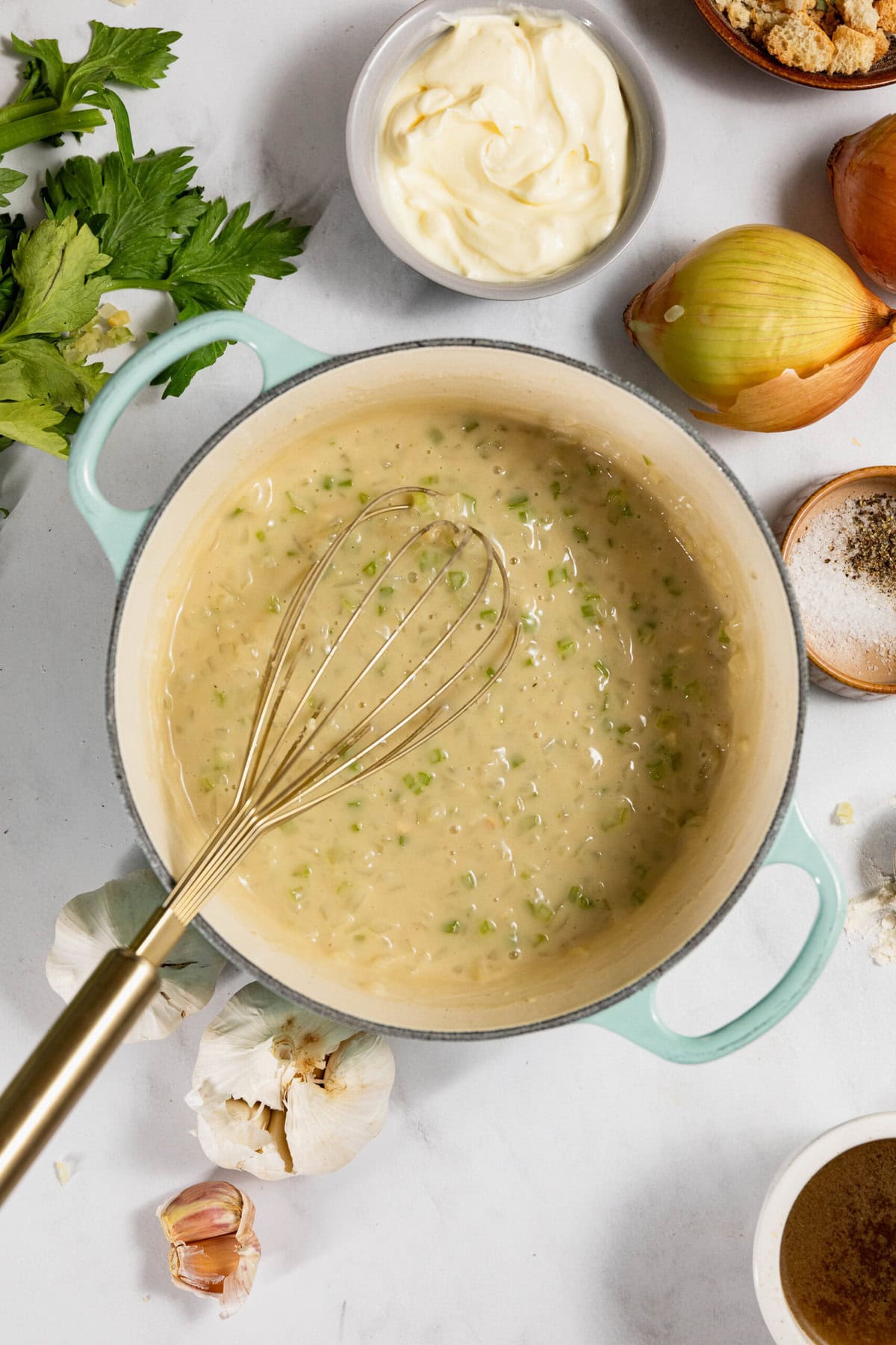 Pot of creamy soup with a whisk, surrounded by ingredients like garlic, onion, celery, mayonnaise, and seasonings on a countertop.