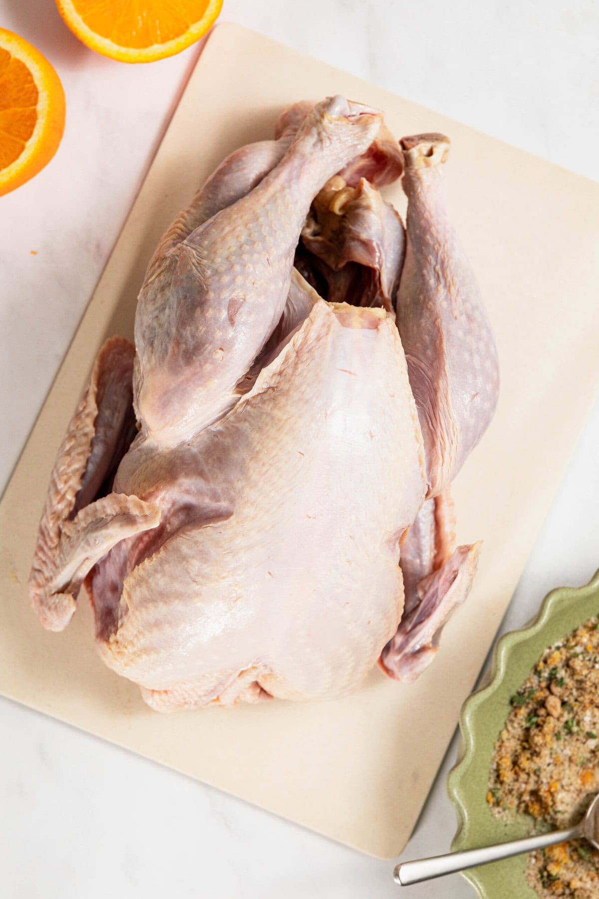 A whole uncooked chicken rests on a cutting board beside two orange halves and a bowl of breadcrumbs with a spoon, reminiscent of preparations for a dry brine turkey.