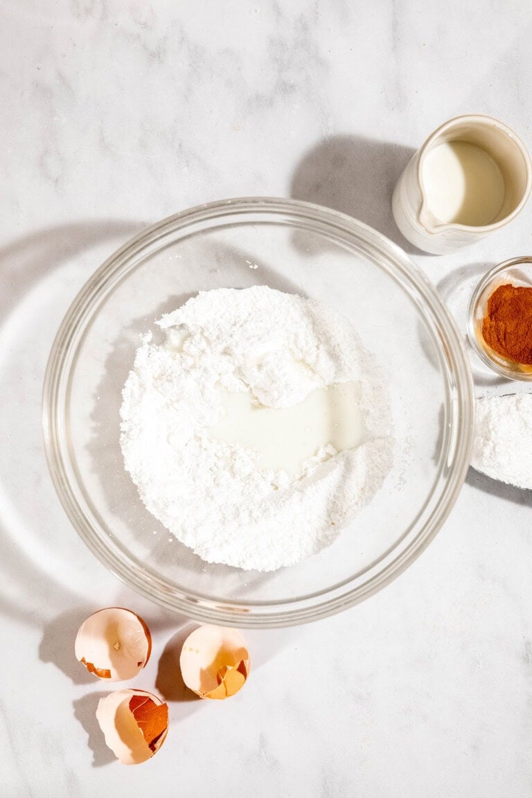 A bowl of flour and milk on a marble surface, surrounded by egg shells, a small jug of milk, a bowl of cinnamon, and a bowl of sugar.