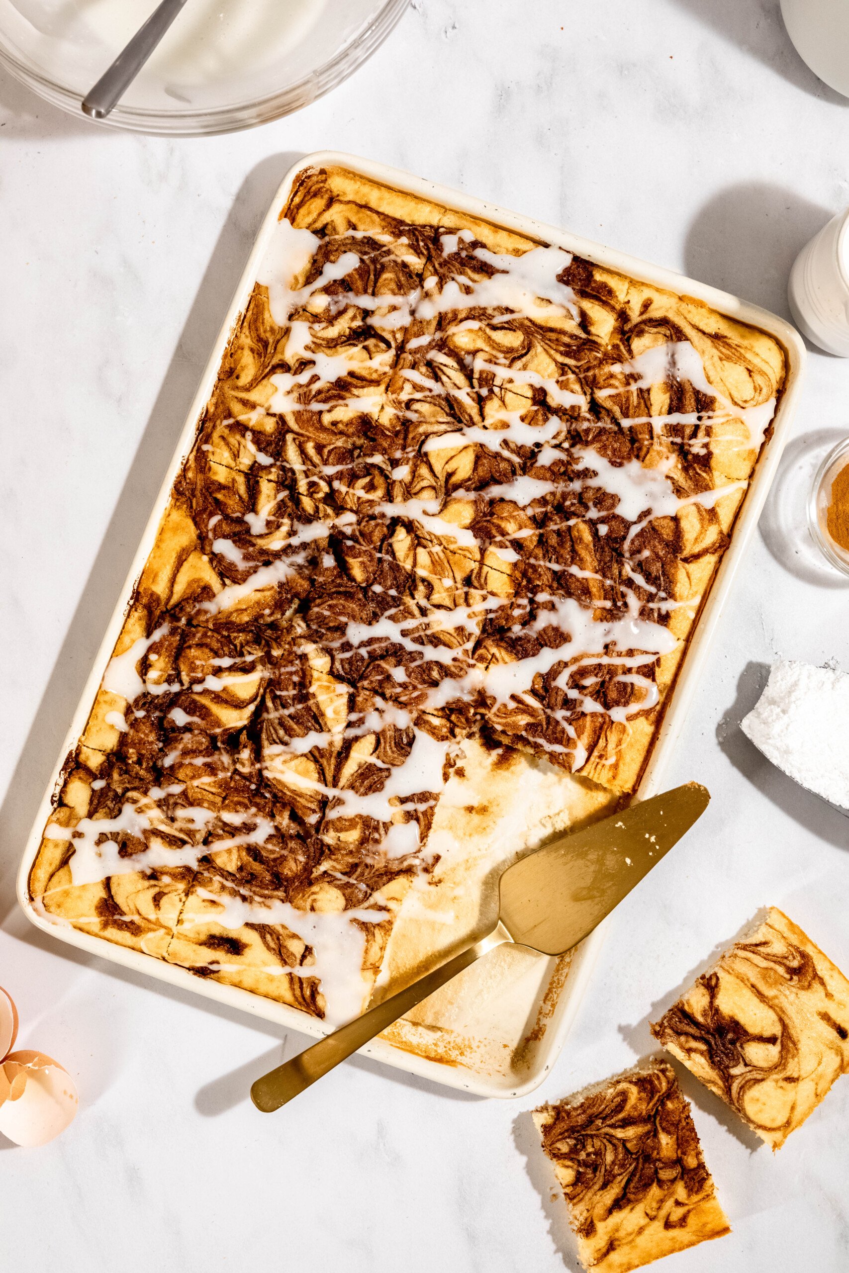 Rectangular marbled cake with icing drizzled on top, partially cut, on a white surface. Golden knife rests beside cake pieces. Nearby are ingredients like flour, eggshells, and seasoning.