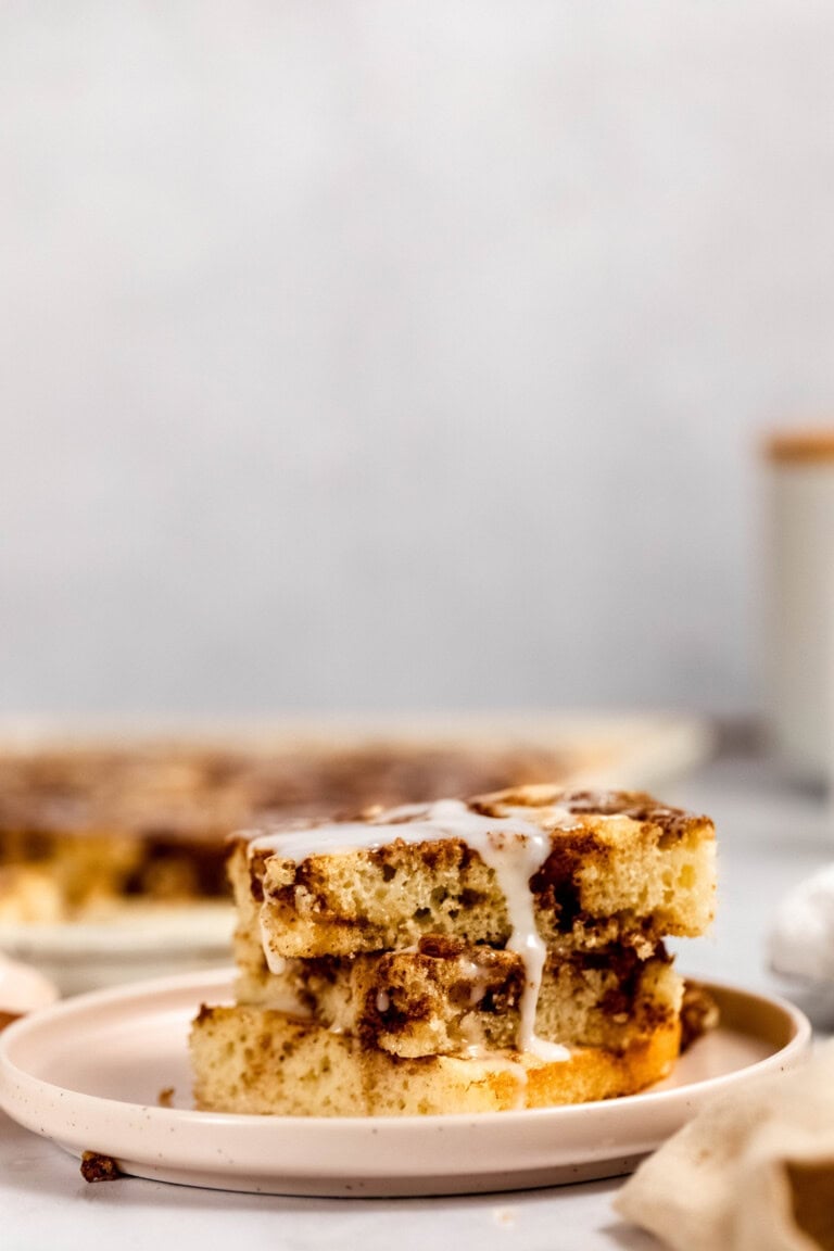 A close-up of three stacked slices of cinnamon coffee cake with icing on a plate. A blurred background shows more cake and a white jar.