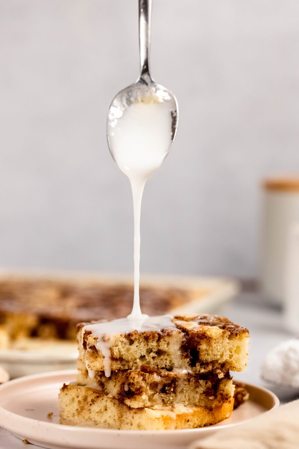 A spoon drizzles white icing over a stack of cinnamon-infused cake squares on a plate.