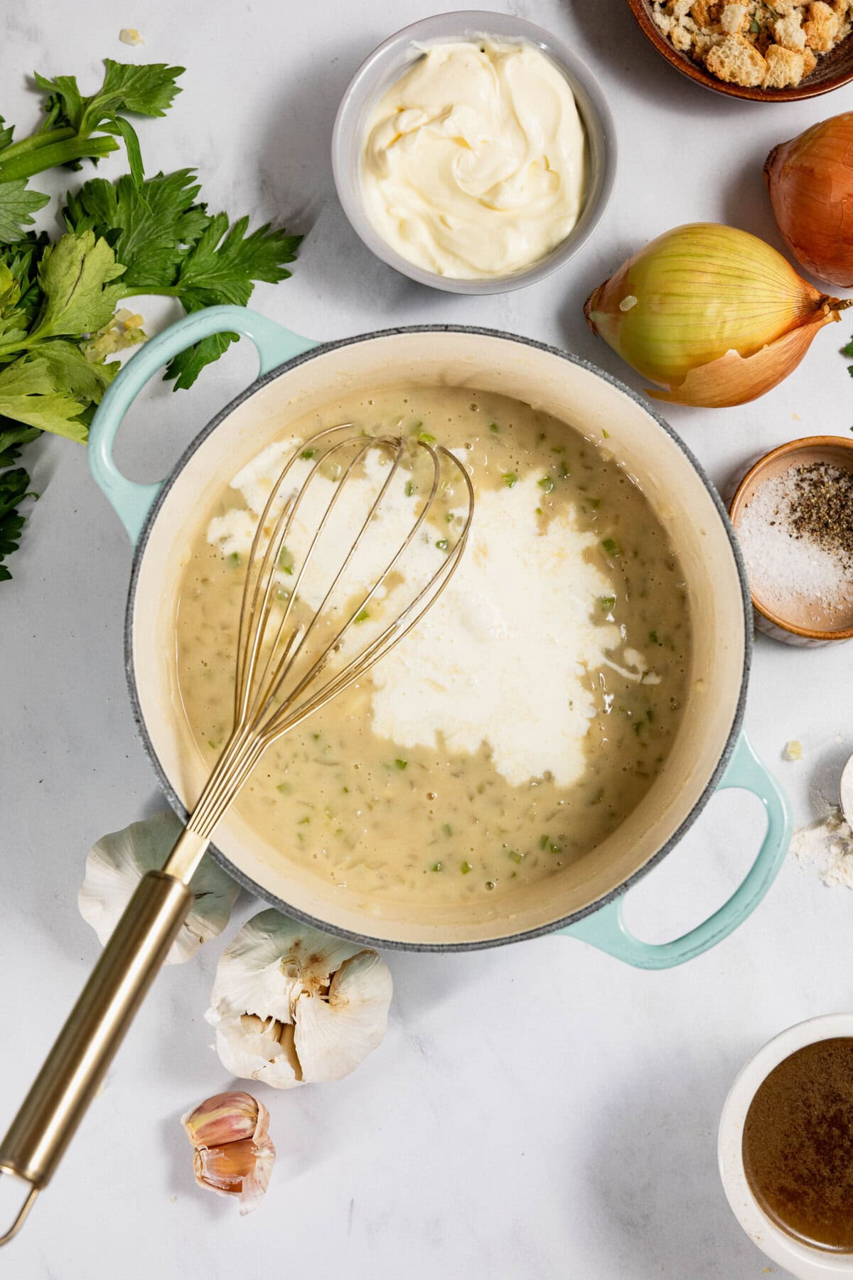 A pot with creamy white soup being whisked, surrounded by ingredients like onions, garlic, bread crumbs, and herbs on a white surface.