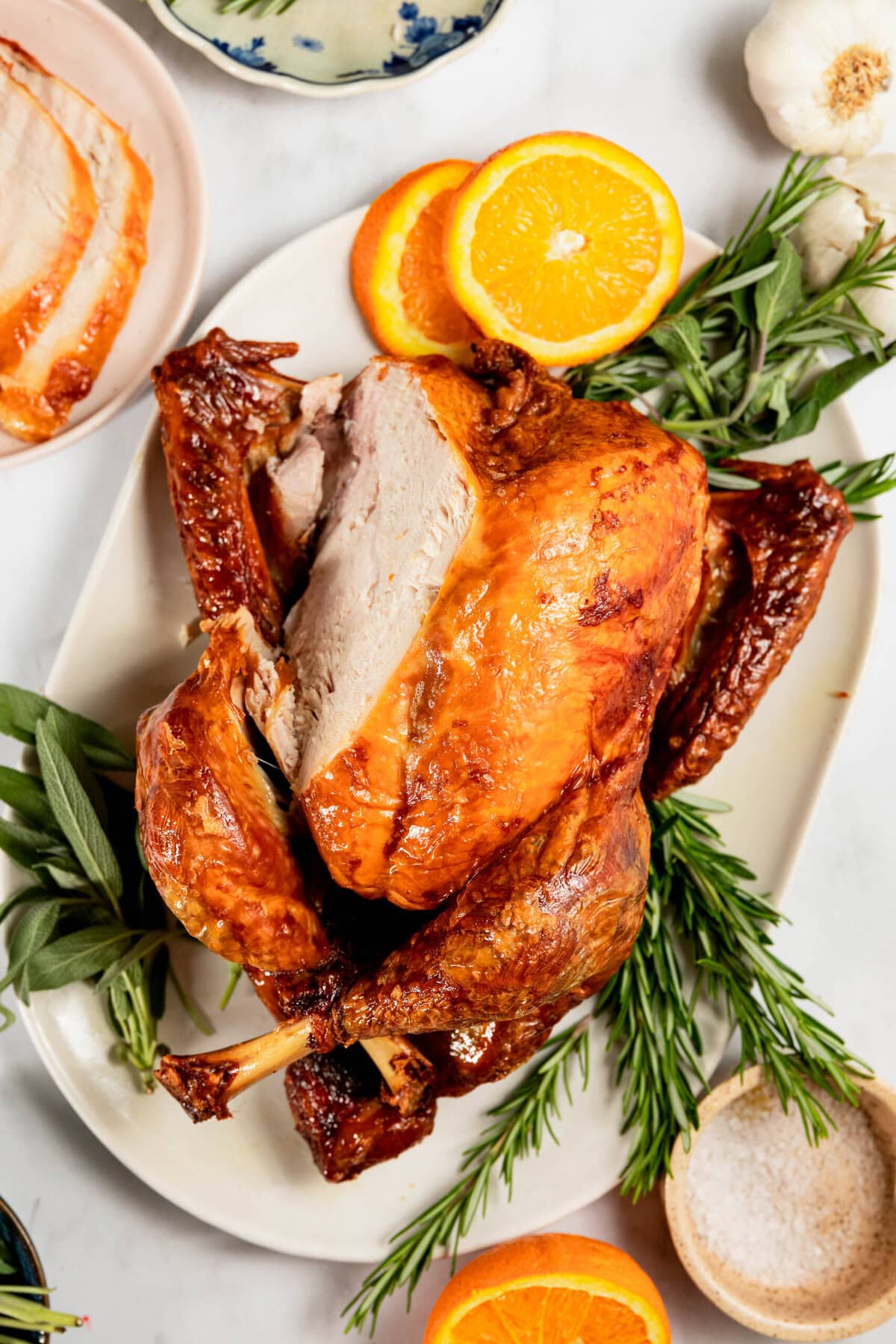 Roasted turkey on a platter garnished with rosemary, sage, and orange slices, surrounded by sliced turkey, oranges, garlic, and a small bowl of salt.