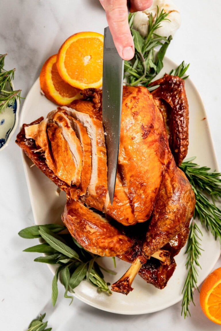 A roasted turkey is being carved on a platter, surrounded by fresh herbs and orange slices.