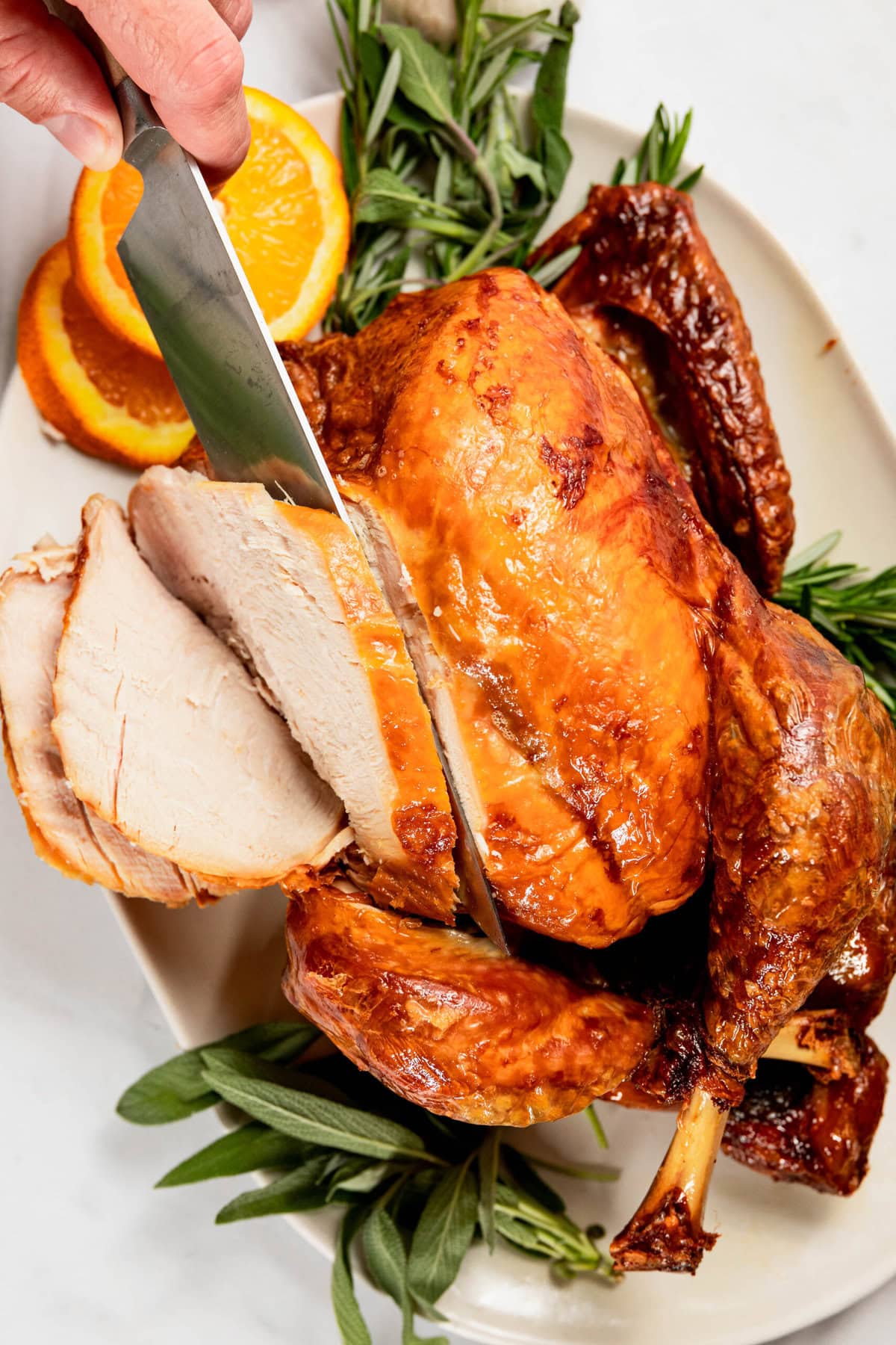 A hand slices a roasted turkey on a platter, garnished with orange slices and herbs.