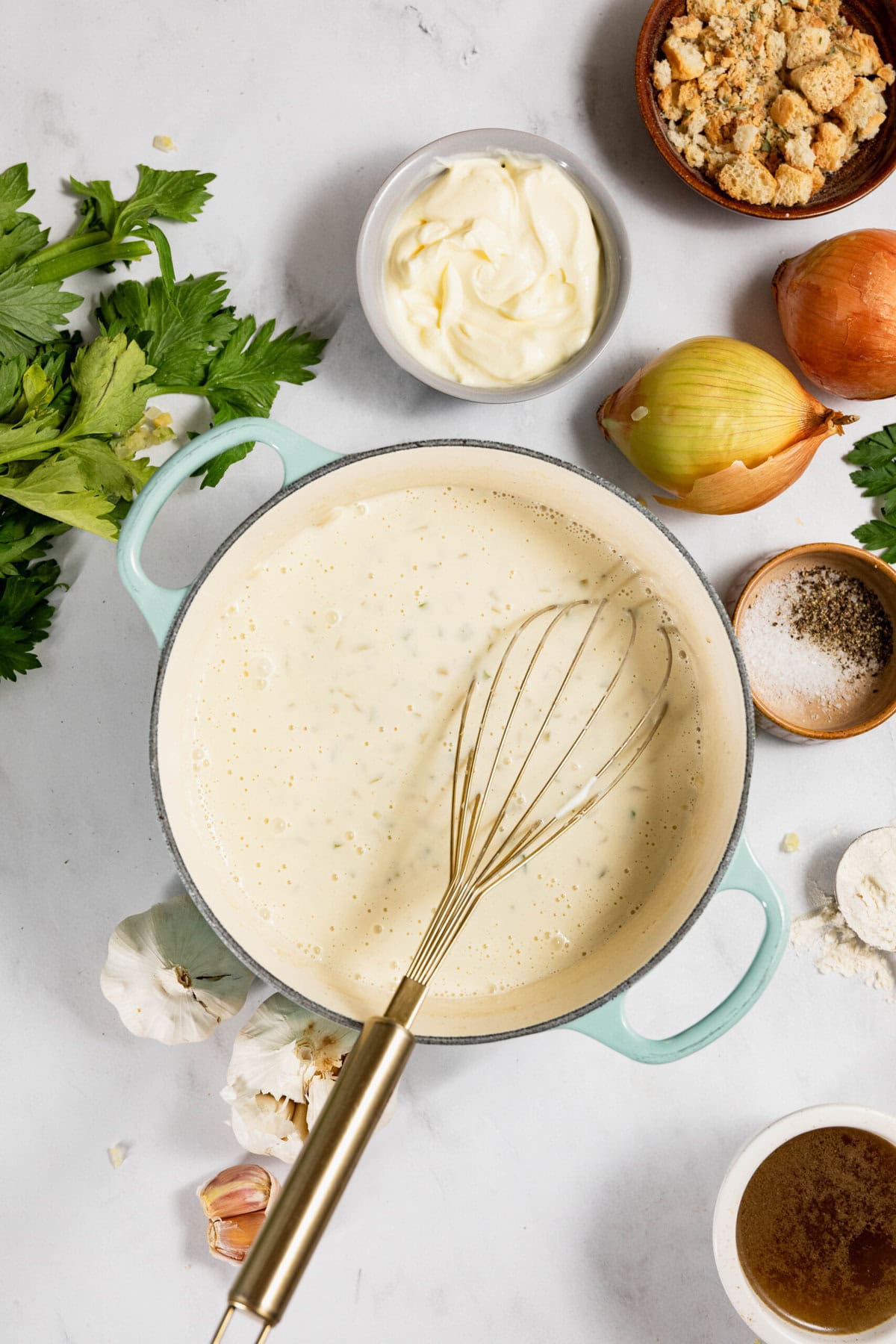 A pot of creamy sauce with a whisk, surrounded by garlic, celery, an onion, breadcrumbs, salt, pepper, flour, and sauce ingredients on a white surface.