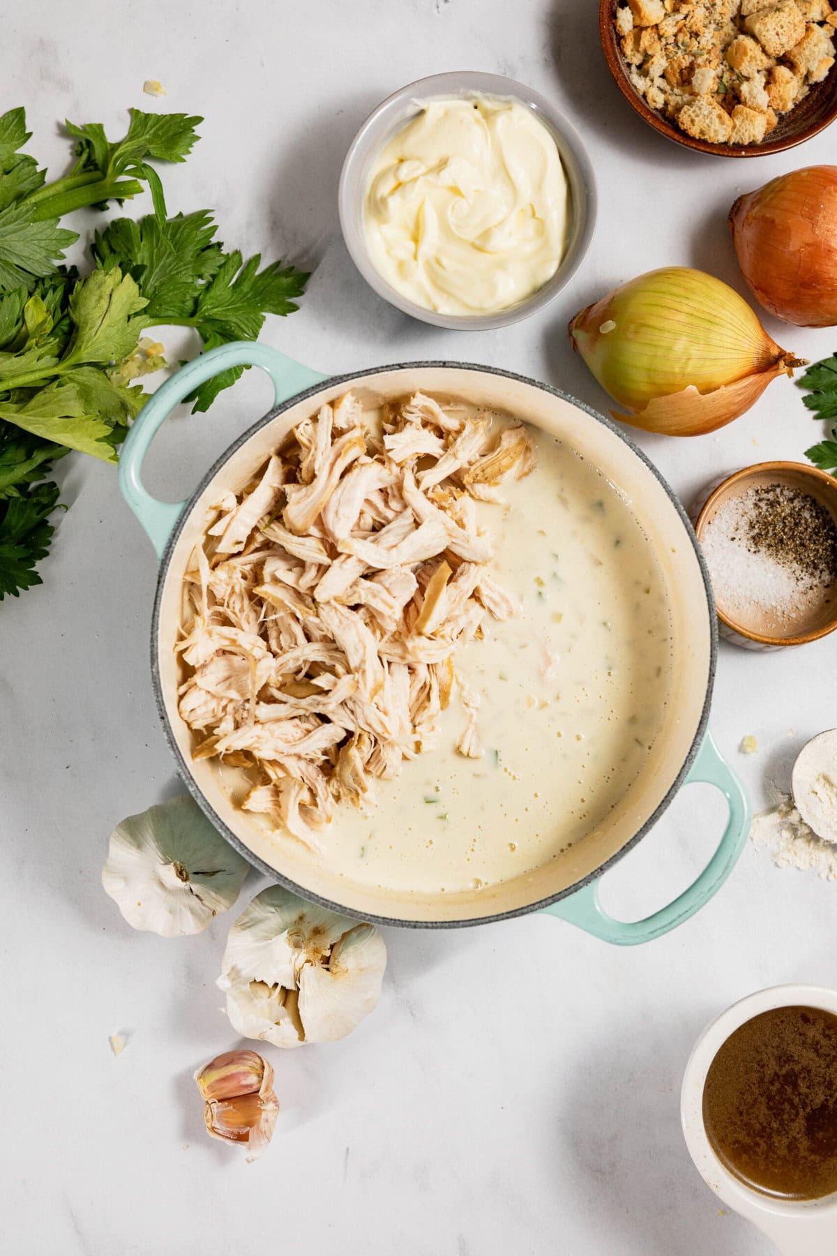 A pot of a creamy mixture with shredded chicken, surrounded by ingredients like onion, garlic, celery, breadcrumbs, and seasoning on a marble surface.