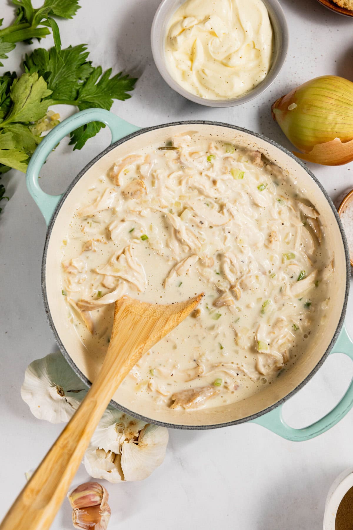 A green pot filled with creamy chicken mixture, stirred with a wooden spoon. Nearby are fresh garlic, an onion, celery leaves, and a bowl of mayonnaise.