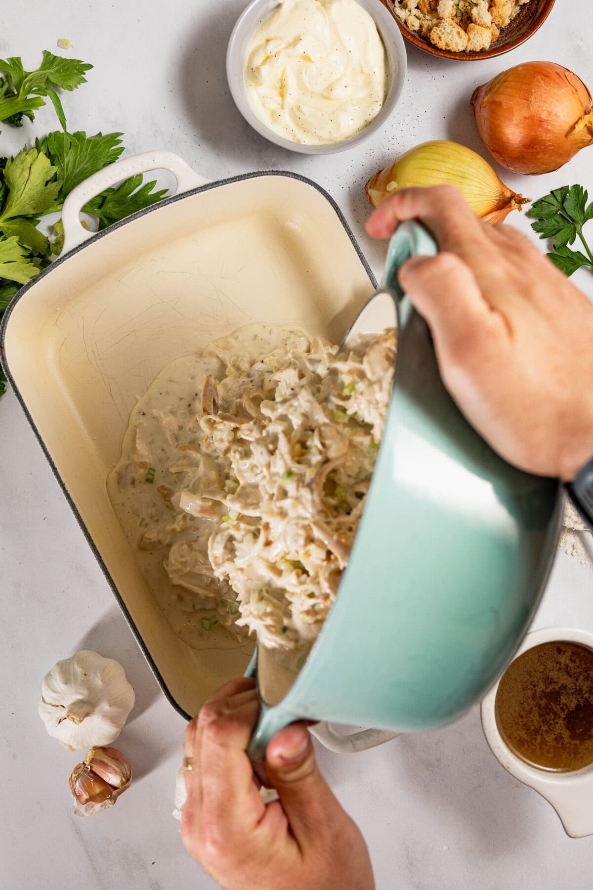 Hands pour a creamy chicken mixture into a baking dish surrounded by ingredients like celery, onions, breadcrumbs, mayonnaise, and garlic on a kitchen countertop.