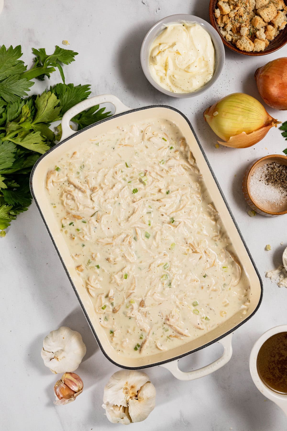 A casserole dish with creamy pasta and shredded chicken, surrounded by ingredients like garlic, onion, spices, and fresh herbs on a gray surface.