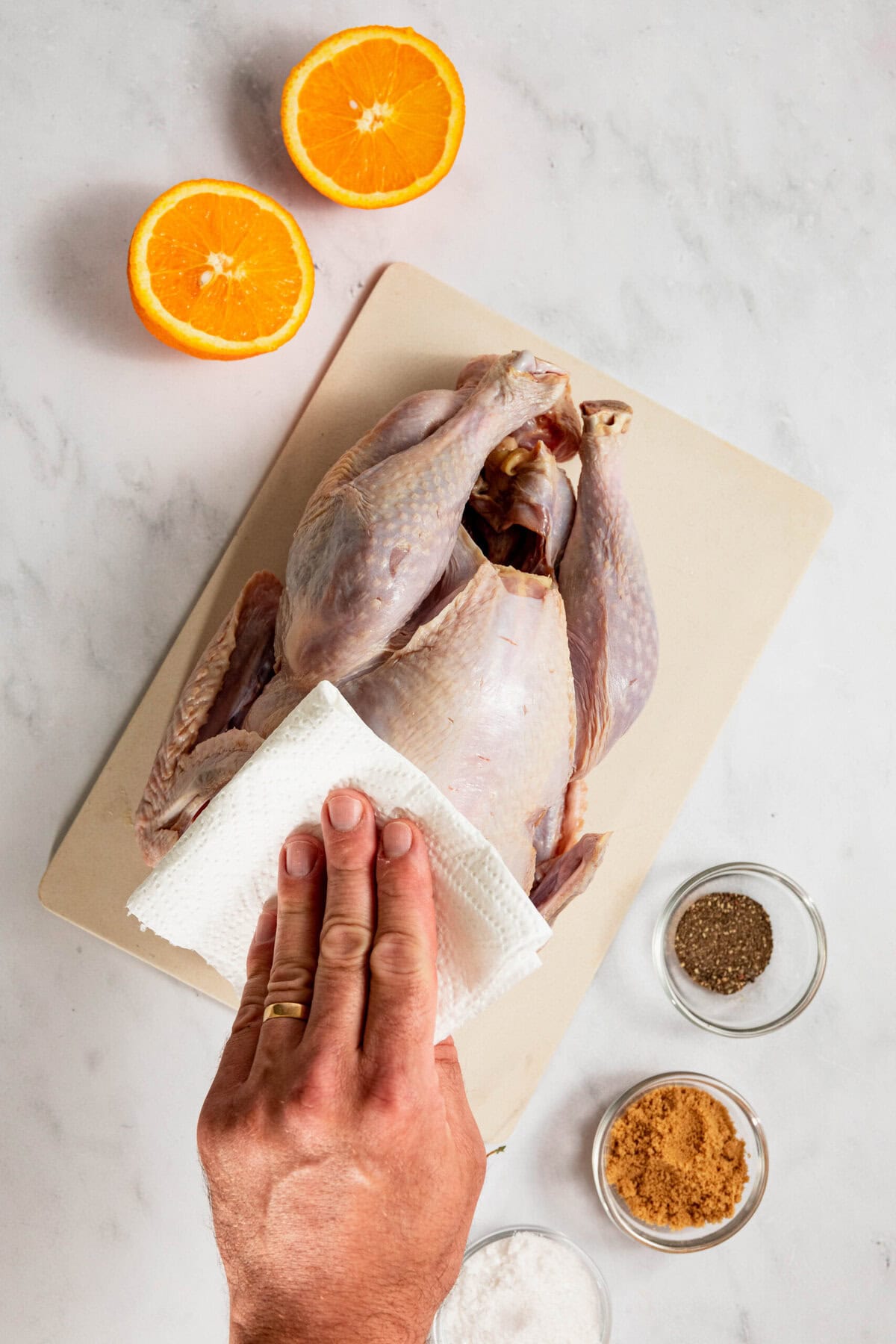 A hand with a ring uses a paper towel to pat a raw chicken on a cutting board, setting the stage for dry brine turkey perfection. Nearby, two halved oranges and three bowls with salt, black pepper, and brown sugar await their flavorful transformation.