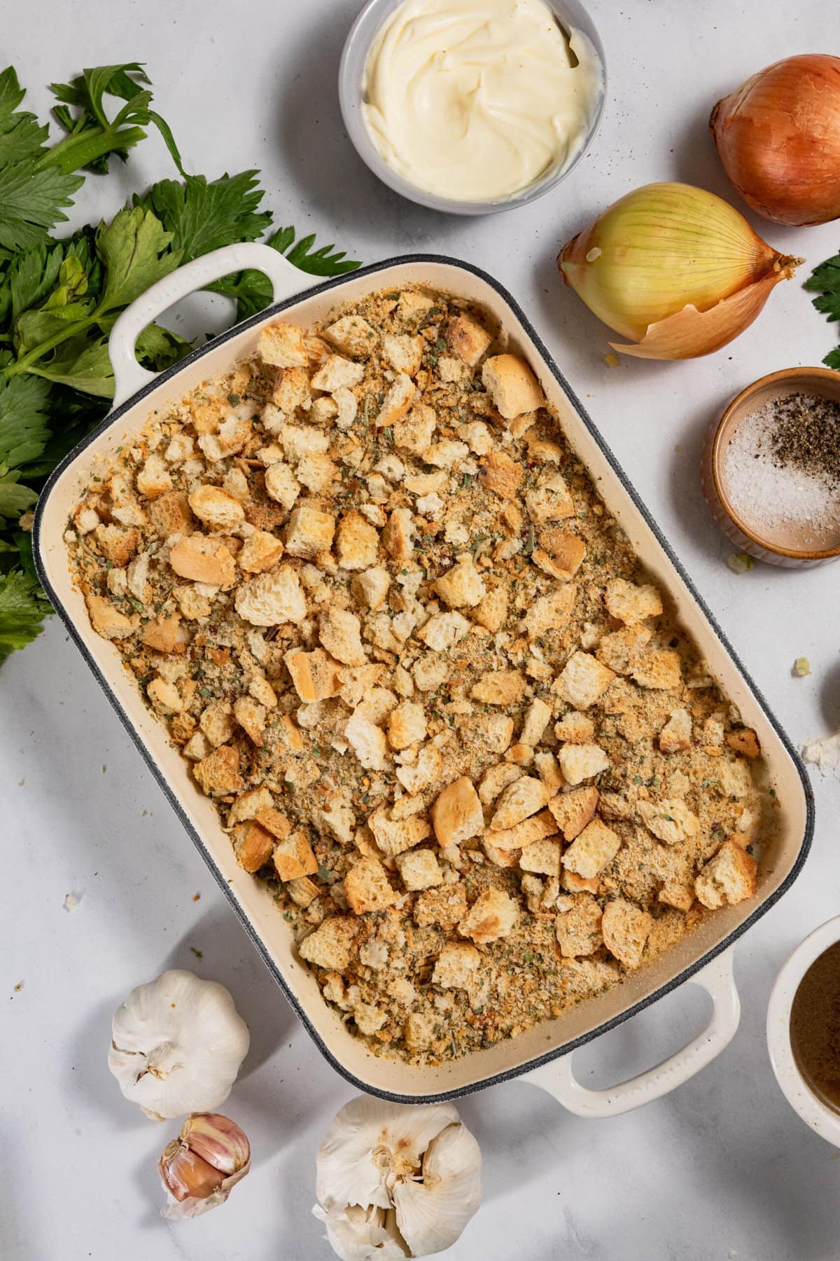 A casserole dish filled with breadcrumb stuffing, surrounded by raw onions, garlic bulbs, fresh parsley, salt, pepper, and a bowl of cream on a white surface.