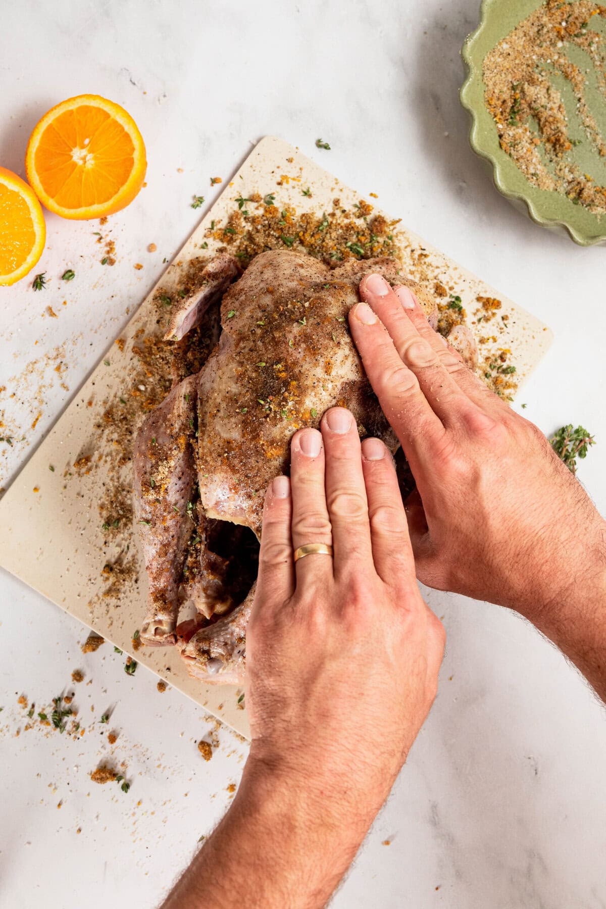 Hands are expertly applying seasoning to a raw chicken on a cutting board, surrounded by vibrant orange halves and a bowl of seasoning. This method, similar to a dry brine turkey technique, enhances the flavor profile for an unforgettable culinary experience.