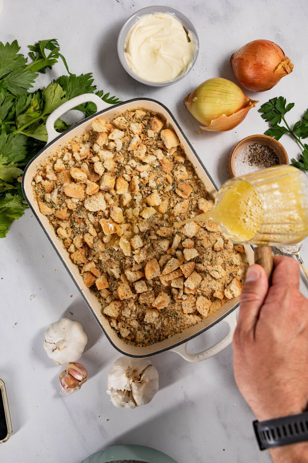 A person pours liquid over a baking dish filled with breadcrumb topping, surrounded by raw ingredients like garlic, onion, green herbs, a bowl of cream, and seasoning on a white countertop.