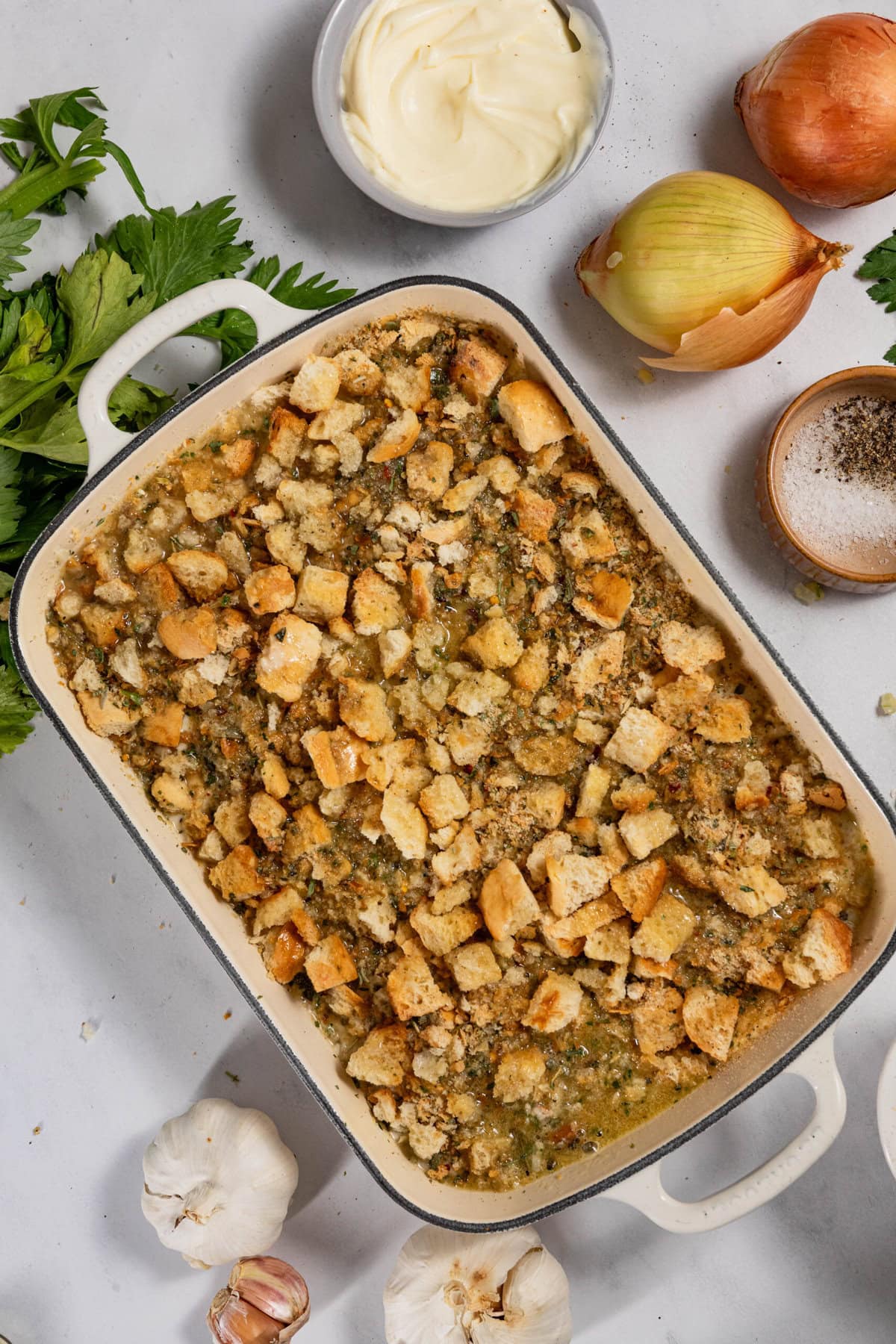 A casserole dish filled with baked stuffing topped with croutons, surrounded by onions, garlic, parsley, a bowl of butter, and small bowls of salt and pepper.