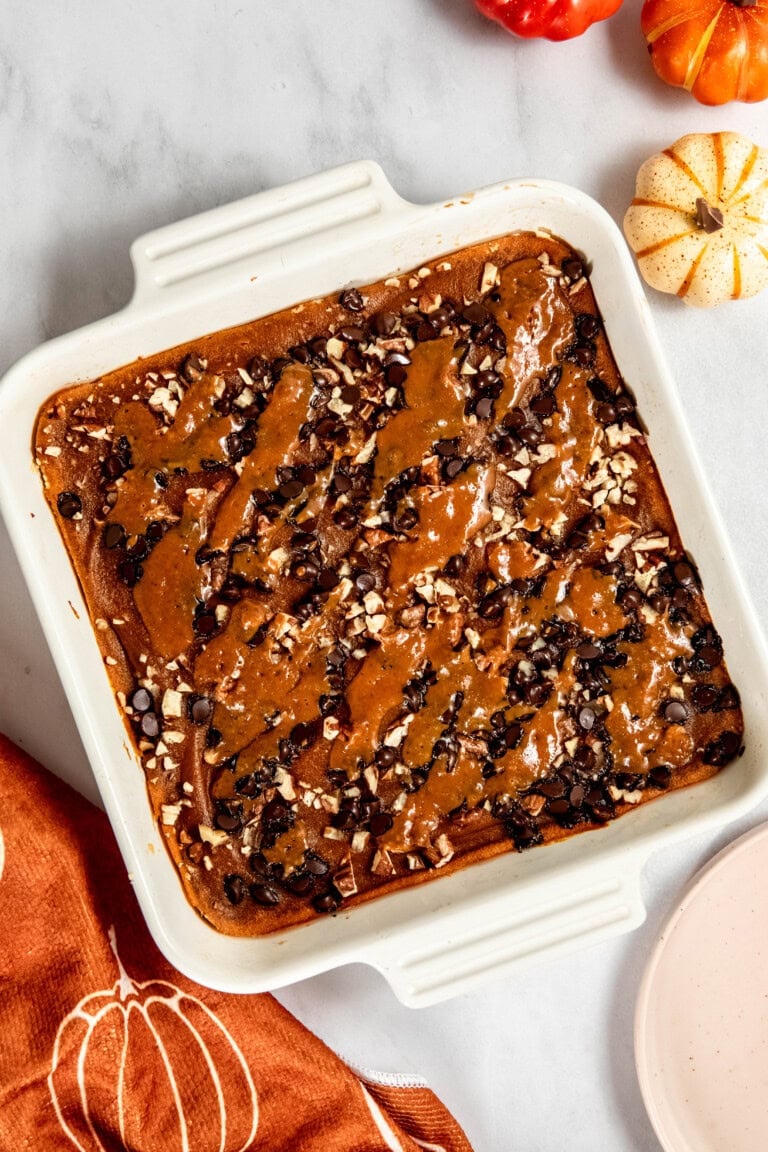 A square baking dish filled with a chocolate dessert topped with nuts and caramel drizzle. Small pumpkins and a pumpkin-themed cloth are nearby.