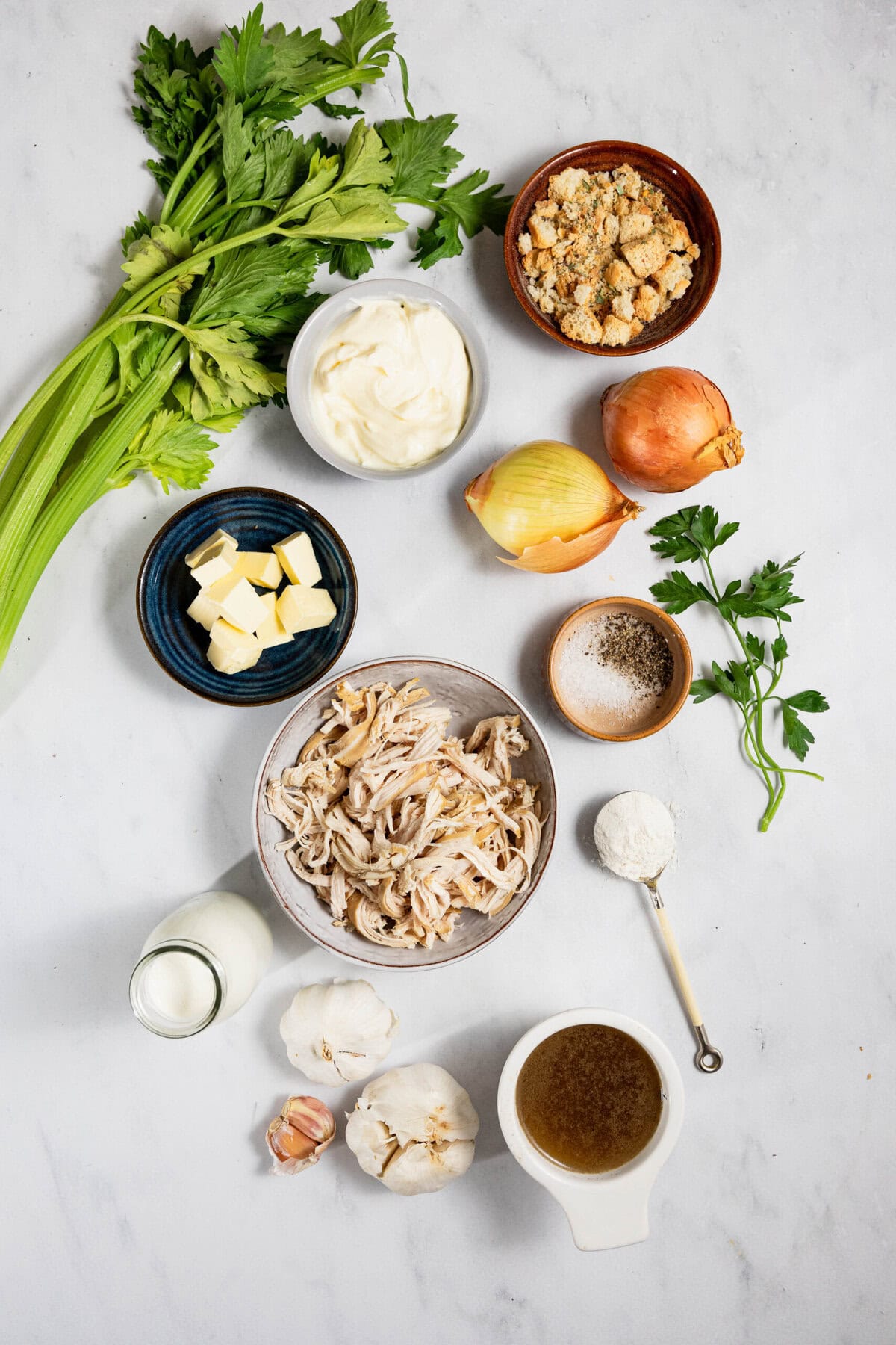 Ingredients for a dish include celery, mayonnaise, bread crumbs, onions, shredded chicken, butter, milk, garlic, broth, parsley, flour, salt, and pepper arranged on a white surface.