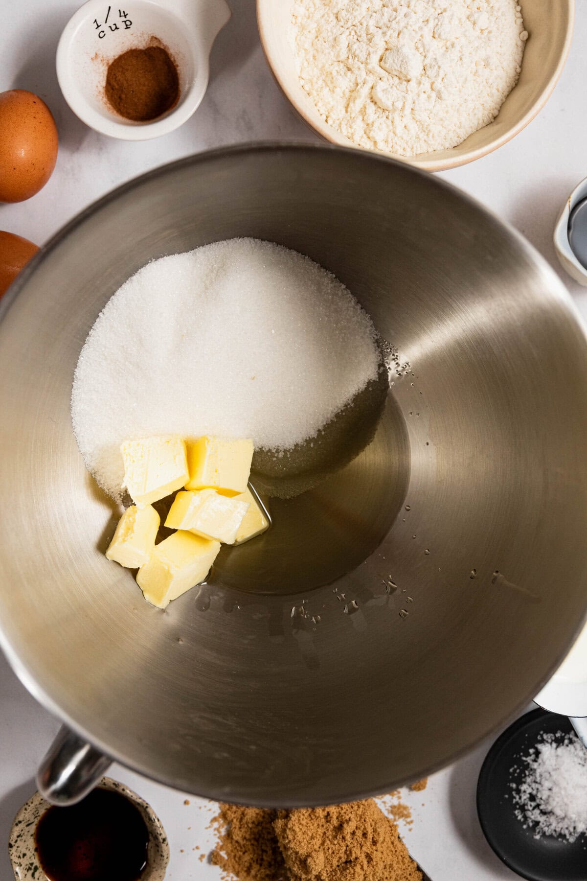 Mixing bowl with sugar and butter, surrounded by flour, eggs, vanilla, cinnamon, brown sugar, and baking ingredients on a countertop.