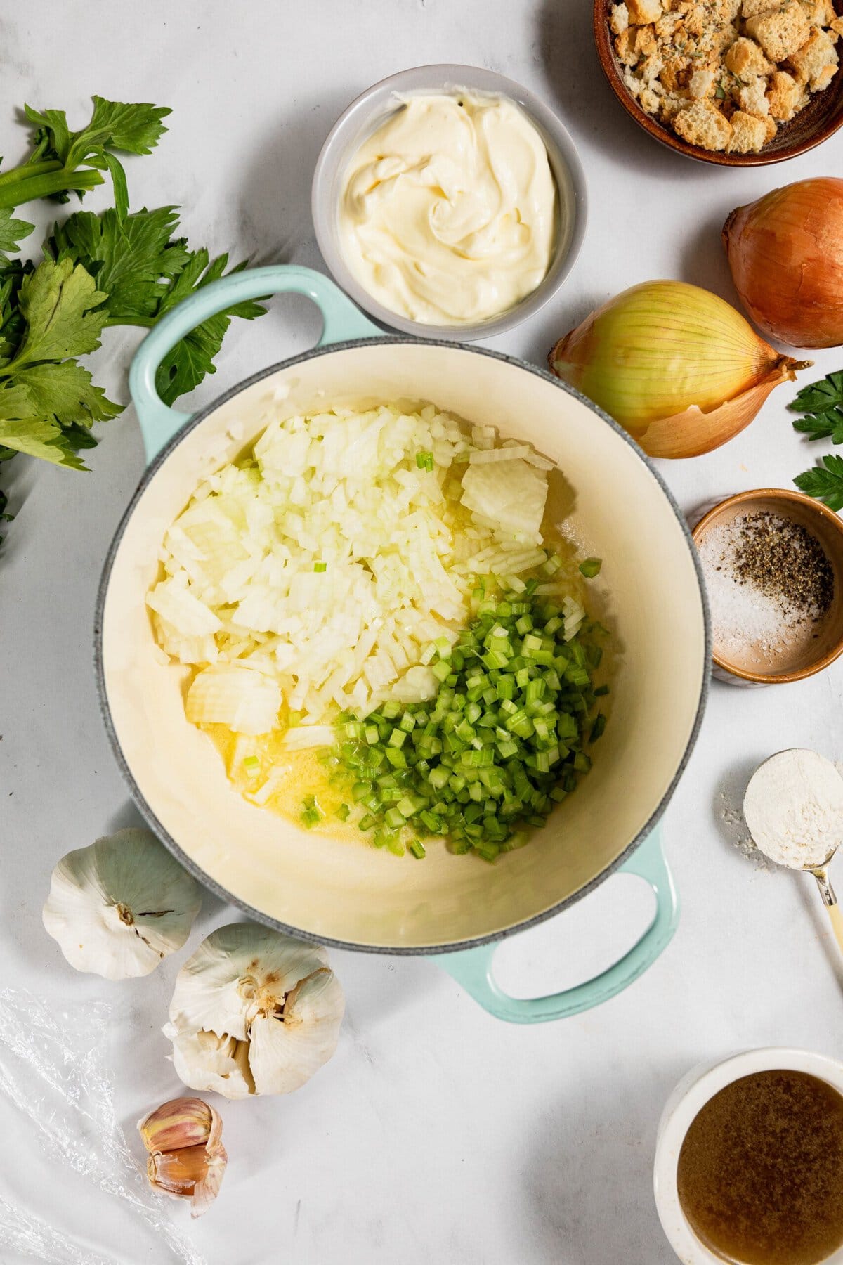 Chopped onions and celery in a pot on a countertop, surrounded by garlic, onion, celery, seasonings, breadcrumbs, and a bowl of mayonnaise.