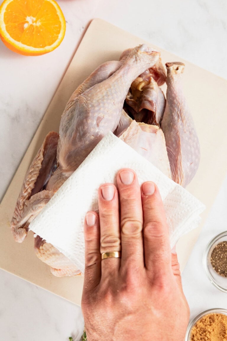 A person uses a paper towel to dry a whole raw chicken on a cutting board, prepping it with the focus and care akin to crafting a perfect dry brine turkey. Ingredients like half an orange and spices are nearby, ready to infuse flavor into the meal.